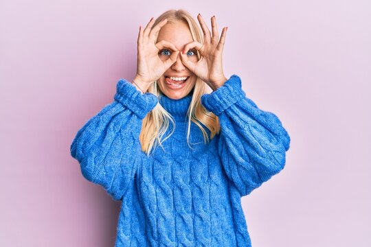 Young blonde girl wearing wool winter sweater doing ok gesture like binoculars sticking tongue out, eyes looking through fingers. crazy expression.