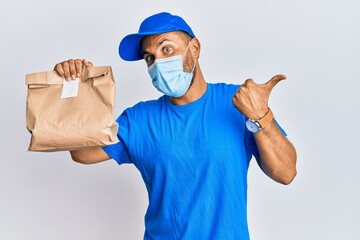 Fototapeta na wymiar Handsome man with beard delivering food wearing covid-19 safety mask pointing thumb up to the side smiling happy with open mouth