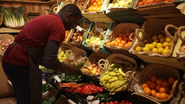 Focused African man working at farmer market, putting fresh fruits and vegetables on showcase