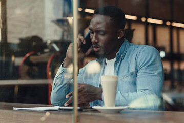 Handsome stylish bearded dark-skinned man sitting near street cafe window and talks on smartphone with business partner.