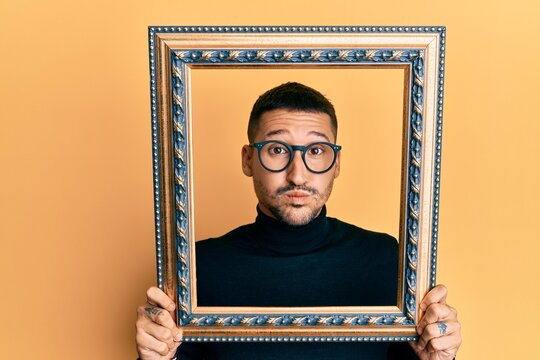 Handsome man with tattoos holding empty frame puffing cheeks with funny face. mouth inflated with air, catching air.