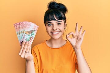 Young hispanic girl holding 100 new zealand dollars banknote doing ok sign with fingers, smiling friendly gesturing excellent symbol