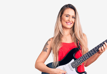 Fototapeta premium Young beautiful blonde woman playing electric guitar looking positive and happy standing and smiling with a confident smile showing teeth