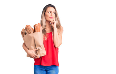 Young beautiful blonde woman holding paper bag with bread serious face thinking about question with...