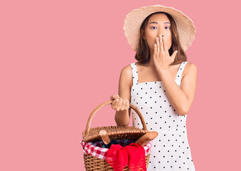 Young beautiful chinese girl wearing summer hat holding picnic basket covering mouth with hand, shocked and afraid for mistake. surprised expression