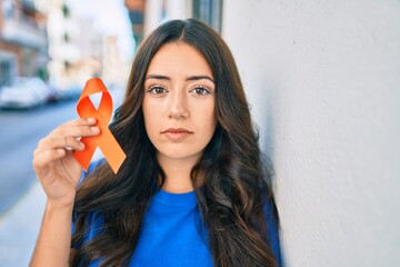 Young hispanic woman with serious expression holding orange ribbon at the city.