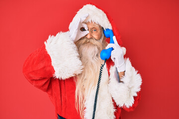 Old senior man wearing santa claus costume using vintage telephone smiling happy doing ok sign with hand on eye looking through fingers