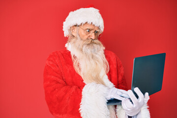 Old senior man with grey hair and long beard wearing santa claus costume using laptop skeptic and nervous, frowning upset because of problem. negative person.