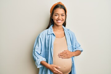 Beautiful hispanic woman expecting a baby, touching pregnant belly with a happy and cool smile on...