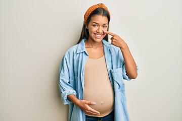 Beautiful hispanic woman expecting a baby, touching pregnant belly pointing with hand finger to face and nose, smiling cheerful. beauty concept
