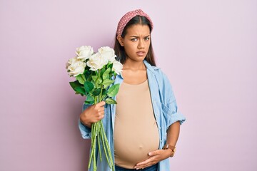 Beautiful hispanic woman expecting a baby holding flowers clueless and confused expression. doubt concept.