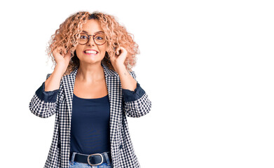Young blonde woman with curly hair wearing business jacket and glasses smiling pulling ears with...