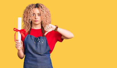 Young blonde woman with curly hair wearing hairdresser apron and holding diploma with angry face, negative sign showing dislike with thumbs down, rejection concept