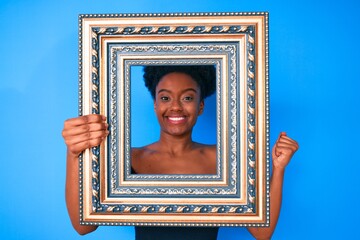 Obraz premium Young african american woman holding empty frame screaming proud, celebrating victory and success very excited with raised arm