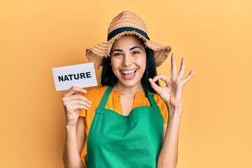 Beautiful young woman wearing gardener apron holding nature word doing ok sign with fingers, smiling friendly gesturing excellent symbol