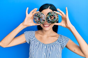 Beautiful young woman holding tasty colorful doughnuts on eyes winking looking at the camera with...