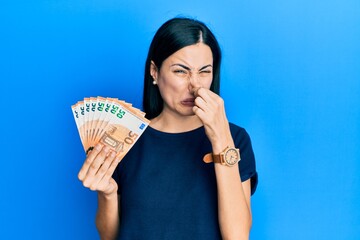 Beautiful young woman holding bunch of 50 euro banknotes smelling something stinky and disgusting,...