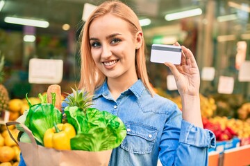 Young blonde girl smiling happy holding groceries paper bag and credit card standing at the supermarket