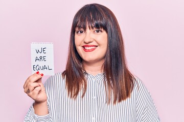 Young plus size woman holding we are equal paper looking positive and happy standing and smiling with a confident smile showing teeth