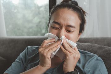 Sick Asian woman sitting indoors holding tissue handkerchief blowing running nose feels unwell unhealthy, girl having symptoms of chronic sinusitis disease, seasonal allergy or cold fever flu concept