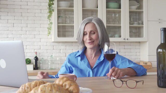 Happy senior woman drinking wine video calling friend on holiday party on laptop at home. Smiling old middle aged lady holding glass talking in online social distance chat sitting at kitchen table.