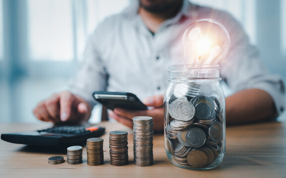 coins stacking on desk and lightbulb in jar with businessman use smartphone and calculator. saving energy and money concept. idea for save or investment.