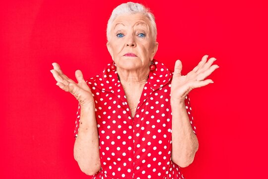 Senior beautiful woman with blue eyes and grey hair wearing casual summer clothes over red background clueless and confused with open arms, no idea and doubtful face.