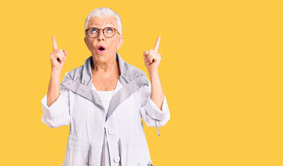 Senior beautiful woman with blue eyes and grey hair wearing casual clothes and glasses amazed and surprised looking up and pointing with fingers and raised arms.