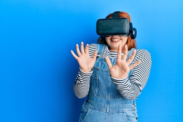 Young redhead woman scared using 3d virtual glasses standing over isolated blue background.