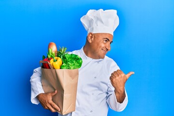 Mature middle east man professional chef holding bag of groceries pointing thumb up to the side smiling happy with open mouth