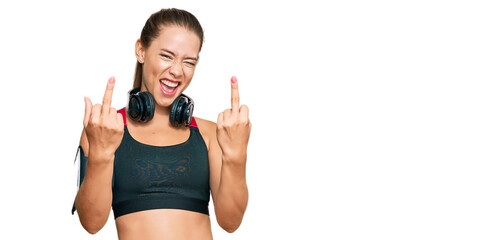 Beautiful blonde woman wearing gym clothes and using headphones showing middle finger doing fuck...