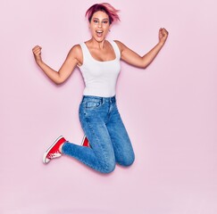 Young beautiful woman with pink short hair wearing casual clothes smiling happy. Jumping with smile on face celebrating with fists up over isolated background.