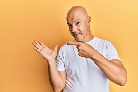 Middle age bald man wearing casual white tshirt amazed and smiling to the camera while presenting with hand and pointing with finger.