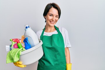 Young brunette woman with short hair wearing apron holding cleaning products winking looking at the...