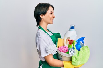 Young brunette woman with short hair wearing apron holding cleaning products looking to side, relax profile pose with natural face and confident smile.