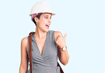 Young beautiful woman wearing architect hardhat and leather bag smiling with happy face looking and pointing to the side with thumb up.