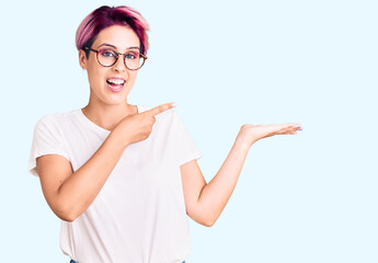 Young beautiful woman with pink hair wearing casual clothes and glasses amazed and smiling to the camera while presenting with hand and pointing with finger.