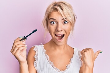 Young blonde girl holding eyelash curler pointing thumb up to the side smiling happy with open mouth