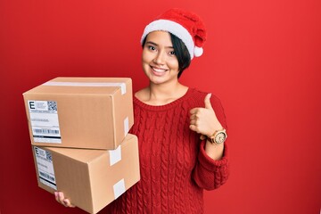 Beautiful young woman with short hair wearing christmas hat holding delivery packages smiling happy and positive, thumb up doing excellent and approval sign