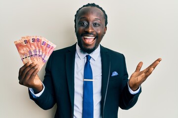 Handsome young black man holding south african rand banknotes celebrating achievement with happy smile and winner expression with raised hand