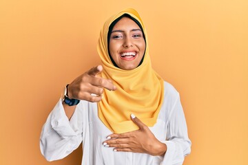 Young brunette arab woman wearing traditional islamic hijab scarf laughing at you, pointing finger to the camera with hand over body, shame expression