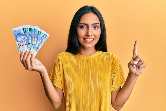 Young brunette woman holding south african 100 rand banknotes smiling with an idea or question pointing finger with happy face, number one