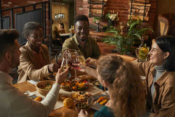 Diverse group of people clinking glasses while enjoying dinner party with friends and family in cozy interior