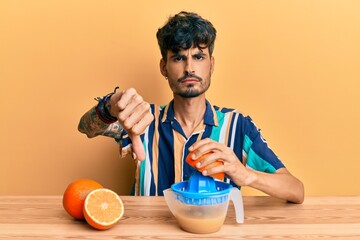 Young hispanic man sitting on the table using juicer with angry face, negative sign showing dislike...