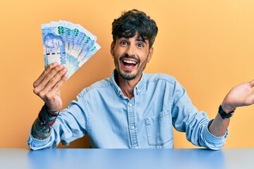 Young hispanic man holding south african rand banknotes sitting on the table celebrating achievement with happy smile and winner expression with raised hand
