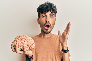 Young hispanic man holding brain scared and amazed with open mouth for surprise, disbelief face