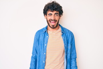 Handsome young man with curly hair and bear wearing casual denim jacket winking looking at the...