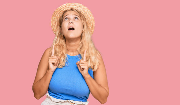 Young blonde girl wearing summer hat amazed and surprised looking up and pointing with fingers and raised arms.