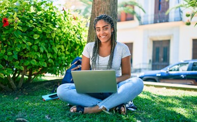 Young african american student woman smiling happy using computer laptop sitting on the grass at the university campus