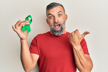 Middle age handsome man holding support green ribbon pointing thumb up to the side smiling happy...